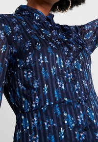 Námořnické šaty na knoflíky s texturovaným pruhovaným vzorem a květinovými akcenty v modré a růžové barvě. Obsahují límec a dlouhé rukávy.