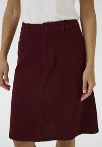 Jupe en corduroy côtelé bordeaux avec une forme évasée, fermeture à boutons sur le devant et poches latérales, présentant un ourlet droit.