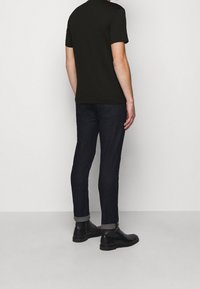 Uomo con una camicia nera a maniche corte, jeans scuri arrotolati e stivali neri in pelle alla caviglia, in piedi contro uno sfondo chiaro e neutro.