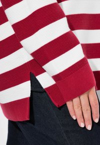Rot-weißer gestreifter Pullover mit gerippter Textur und seitlichen Schlitzen, getragen über dunkelblauen Jeans, die eine lässige Passform und entspanntes Design zeigen.