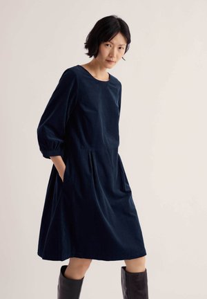 Femme aux cheveux courts noirs portant une robe ample bleu marine avec des poches et des bottes noires montant jusqu'aux genoux, debout devant un fond clair uni.