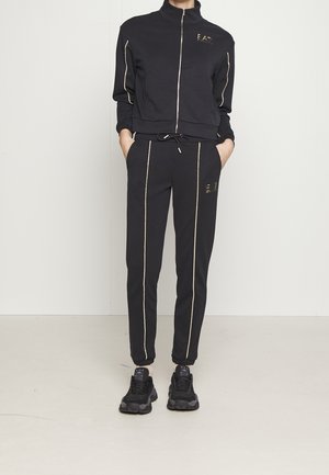 Personne portant une veste noire zippée et un pantalon assorti avec des rayures verticales dorées et des baskets noires épaisses, mains dans les poches.