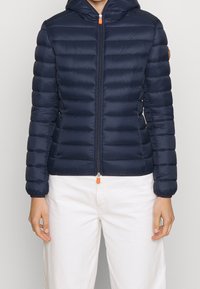 Mørkeblå pufjakke med hætte, der har vandret quiltet design, frontlukning med lynlås og orange lynlåsdetalje. Blød tekstur.