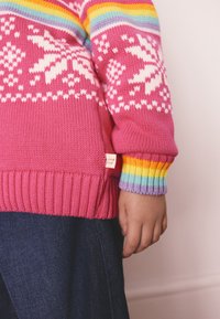 Neulottu kirkkaanpunainen villapaita, jossa valkoiset lumihiutalekuvioinnit, resoriset mansetit, joissa sateenkaariraidat, ja pieni etiketti helmassa.