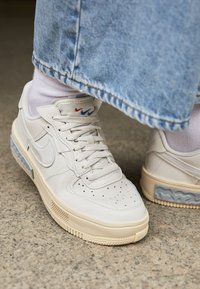 Beżowy skórzany sneaker z perforowanym przodem, gumową podeszwą oraz logo w kształcie trójkolorowego znaku. W towarzystwie jasnoniebieskiego mankietu spodni dżinsowych.