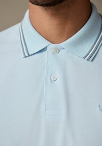 Svetle modrá polo tričko s texturovanou vazbou, dvěma knoflíky a límečkem s bílými pruhy. Diskrétní logo vyšívané na hrudi.