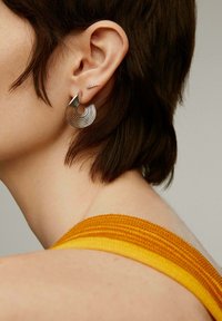 Pendiente de aro en espiral de plata con anillos concéntricos; pequeño pendiente de botón de plata con un sutil detalle de gema. Modelo llevando una blusa amarilla de tirantes.