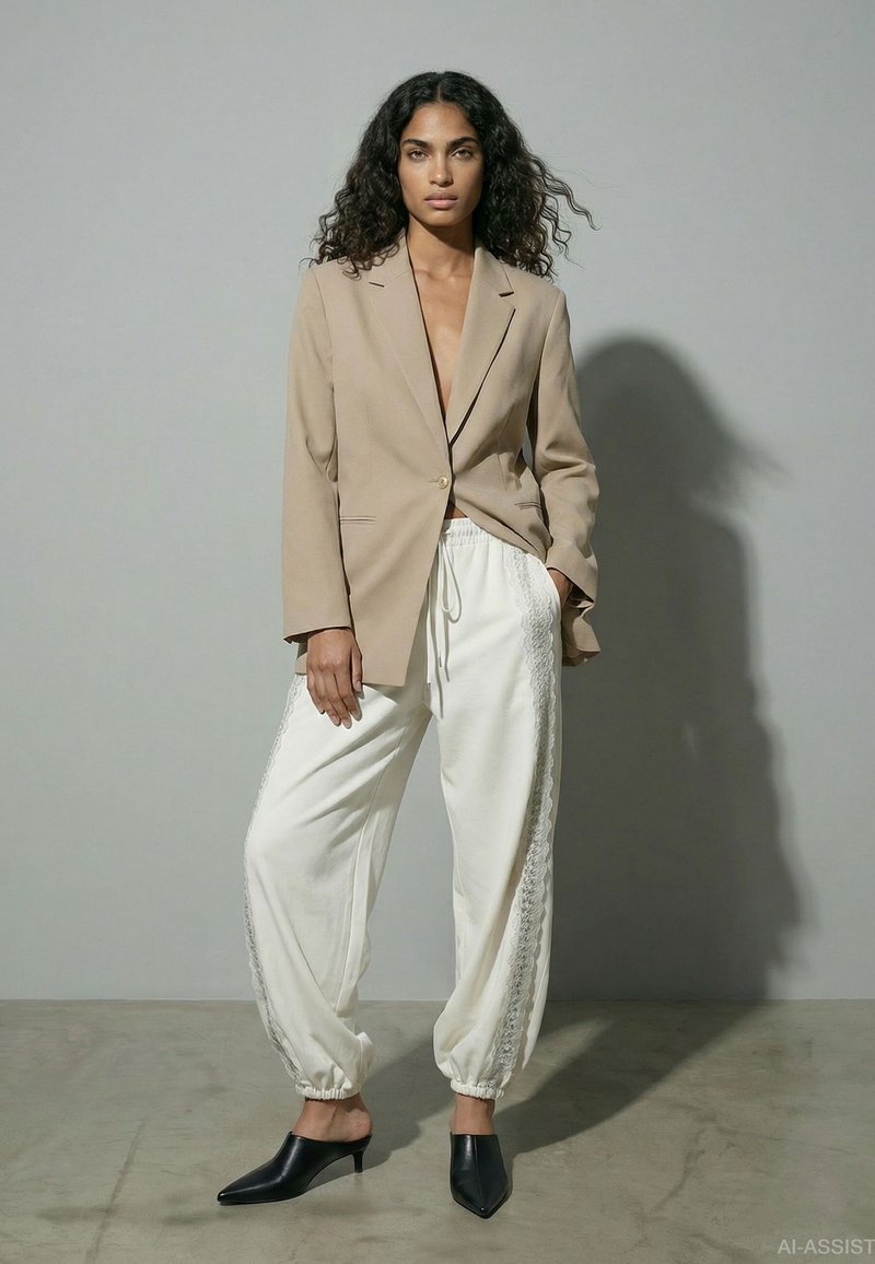 Femme aux cheveux bouclés portant un blazer beige à un bouton, un pantalon blanc à cordon avec des côtés en dentelle et des mules noires à talons, debout contre un mur gris.