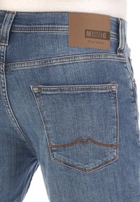 Niebieskie dżinsy z denimu z kontrastowymi pomarańczowymi szwami, tylną kieszenią o ostrym kształcie oraz brązową skórzaną metką z napisem "MUSTANG True Denim".