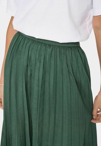 Gonna verde plissettata con finitura testurizzata. La vita è elastica e il tessuto ha una leggera lucentezza. Accompagnata da una maglietta bianca semplice.
