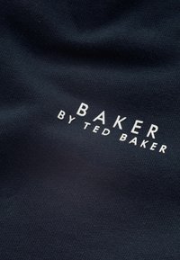 Granatowy materiał o gładkiej teksturze z białym napisem "BAKER", po którym znajduje się "by TED BAKER," umieszczony centralnie.