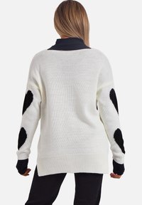 Cremefarbenes Strickpullover mit großen schwarzen Herzmustern an den Ärmeln, gerippten Bündchen und einem leicht verlängerten Rückensaum.