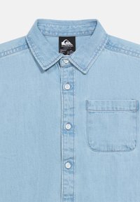 Ανοιχτό μπλε πουκάμισο από τζιν με κουμπιά, γιακά και τσέπη στο στήθος, με λευκά κουμπιά και ορατή μαύρη ετικέτα της μάρκας μέσα στον γιακά.