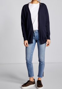 Marinblå cardigan, lätt stickad, öppen framsidestil. Vit skjorta under, ljusblå jeans och mönstrade bruna sneakers. Croppad passform med rå nederkant.