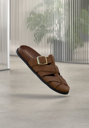 Sandale en cuir marron avec boucle dorée et semelle noire, flottant au-dessus d'un sol en béton lisse, avec en arrière-plan un glass rayé verticalement.