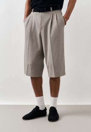 Férfi, aki bézs és fekete kockás, térdig érő, pliszírozott rövidnadrágot visel, fekete övvel, fehér zoknival, fekete sportcipővel és fekete inggel, kezek a zsebekben.