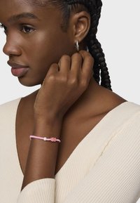 Bracciale intrecciato rosa con una piccola perla e un fermaglio decorativo rosa, indossato su una mano di carnagione scura su uno sfondo chiaro.