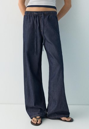 Persoană purtând pantaloni evazați din denim albastru închis, cu talie cu șnur și papuci negri, stând pe un fundal simplu, deschis.