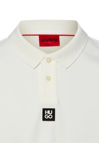 Camiseta tipo polo blanca con textura suave, tapeta de tres botones y un parche cuadrado negro con la marca "HUGO" en blanco. Detalle en rojo en el interior del cuello.