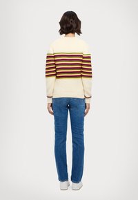 Pull en crème avec des rayures horizontales colorées en rouge, jaune, vert et violet ; poignets et ourlet côtelés ; porté avec un jean bleu.