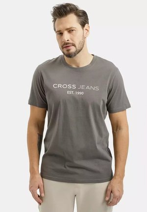 Mężczyzna z krótkimi włosami i brodą, ubrany w szarą koszulkę z napisem „CROSS JEANS EST. 1990” oraz jasnobeżowe spodnie, stojący na jednolitym tle.
