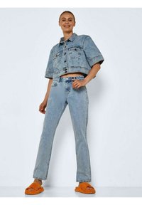 Jaqueta de denim azul claro, cropped, com mangas curtas, combinada com jeans azul claro de perna reta. Usada com sandálias texturizadas laranja brilhante.