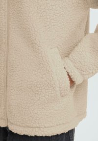 Kreemikas fliisjakk tekstuurse, kohevusega pinnaga, millel on küljetaskud ja lukk. Serv on veidi kõverdatud.