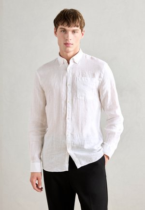 Cămașă albă din in cu mâneci lungi, închidere cu nasturi, un buzunar pe piept și un tiv rotunjit. Asortată cu pantaloni negri, pe un fundal neutru.