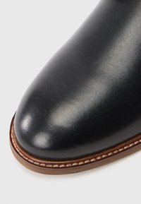 Chaussure en cuir noir avec un bout rond, texture lisse et une couture marron contrastante le long de la semelle pour plus de détails.