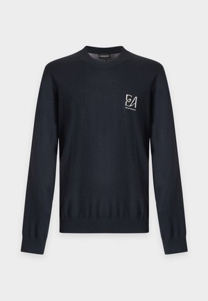 Homme portant une chemise noire à manches longues, un pantalon marron et des mocassins noirs, debout les mains dans les poches sur fond blanc.