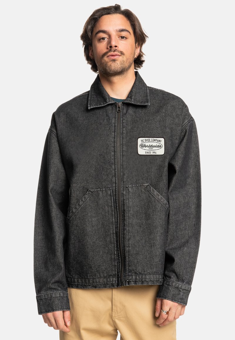 Black denim jacket with a front zipper, two side pockets, and a logo patch on the left chest. Features a classic collar and relaxed fit.