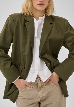Blonde vrouw met een olijfgroene blazer over een wit overhemd en beige broek, met handen in de zakken tegen een effen achtergrond.