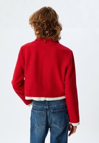 Rote, gekürzte Jacke aus strukturiertem Stoff, mit Spitzkragen und langen Ärmeln. Über blauen Jeans und einem weißen Unterhemd am Saum getragen.