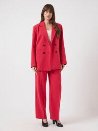 Kvinne som står iført en lys rød oversized dobbeltradet blazer og matchende vide bukser, med svarte hæler og hvit topp.