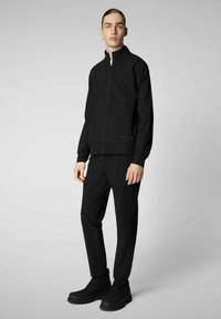 Giacca nera con zip e collo alto, abbinata a pantaloni neri e stivali. Tessuto liscio, design minimalista e vestibilità su misura.