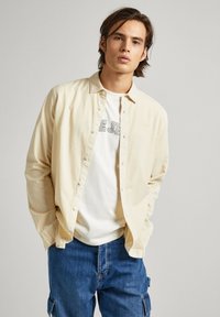 Camicia button-up beige chiaro con righe verticali, indossata sopra una maglietta bianca con scritte nere, abbinata a pantaloni cargo in denim blu.