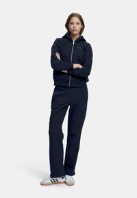 Marineblå hettegenser med glidelås og ribbestrikkede mansjetter, parret med matchende joggebukser; begge laget av myk stoff, med subtile merker.