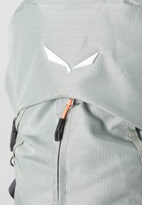 Light gray backpack made of textured fabric with a white logo detail and an orange zipper pull for easy access. Features adjustable straps.