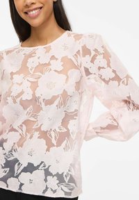 Femme portant un chemisier transparent rose pâle avec de grandes broderies florales blanches et des manches longues bouffantes, souriant doucement.