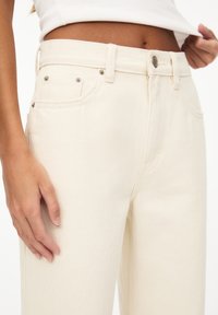 Pantalon en denim couleur crème avec taille haute, design à cinq poches et accents métalliques sur la ceinture. Texture chevrons visible.