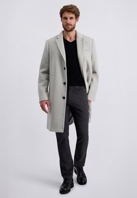 Man loopt vooruit in een lichtgrijze overjas, zwart V-hals shirt, donkergrijze broek en zwarte veterschoenen tegen een effen achtergrond.