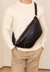 Schwarze Leder-Schultertasche mit einer geschwungenen Form, ausgestattet mit einem goldenen Reißverschluss und einem verstellbaren Gurt, getragen über einem beigen Pullover und einer Hose.