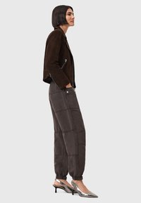 Giacca in suede marrone, pantaloni grigi a vestibilità rilassata con pannelli cuciti e décolleté slingback metallici con punta affusolata. Sfondo neutro.