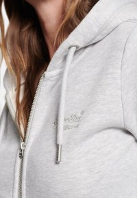 Sudadera con capucha gris con cremallera y cordones, con textura suave y logo bordado en el pecho. Cremallera plateada y extremos metálicos en los cordones.