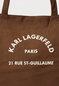 Geantă tote maro din pânză, cu text alb care scrie „KARL LAGERFELD PARIS 21 RUE ST-GUILLAUME” într-un font modern, cu un design curbat.