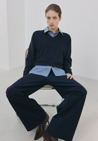 Kvinde sidder på stol med benene spredt, iført mørke bukser med brede ben, navyblå trøje over blåstribet skjorte og brune spidse støvler.