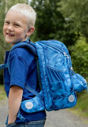 Smiling young boy wearing a blue shirt and large blue geometric-patterned backpack outdoors on a dirt path with greenery in the background.