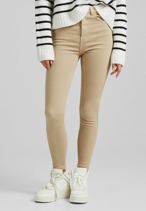 Persona vistiendo pantalones ajustados beige, zapatillas blancas y un suéter blanco con rayas horizontales negras, de pie frente a un fondo gris liso.