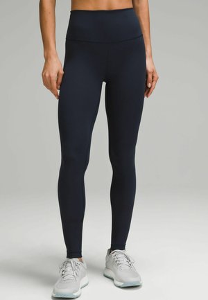 Marineblaue Leggings mit hohem Bund aus glattem, dehnbarem Stoff. Das eng anliegende Design reicht bis zu den Knöcheln. Kombiniert mit grauen Sportschuhen.