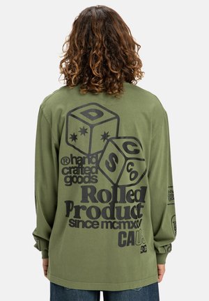 Persoon met krullend haar draagt een olijfgroene longsleeve met grote zwarte grafische dobbelstenen en de tekst "handcrafted goods Rolled Product" op de achterkant.